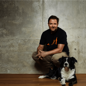Hockender Martin Rütter befindet sich vor einer Wand mit einem liegenden Hund 