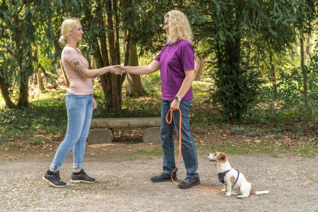 Zwei Frauen schütteln sich die Hand während der Hund neben der Halterin sitzt und wartet.